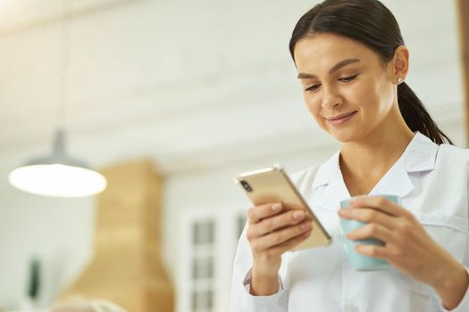 Smiling young nurse holding cup and looking at mobile phone screen while standing in room