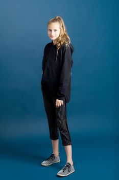 Teenage girl in black hoodie and plaid trousers. Full length shot of beautiful girl dressed sweatshirt, breeches and sneakers posing against blue background in studio looking seriously at camera