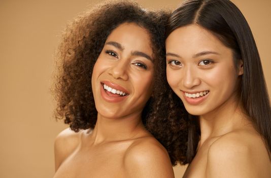 Portrait of two joyful attractive mixed race young women with perfect smile posing for camera isolated over beige background. Health and beauty, diversity concept. Selective focus
