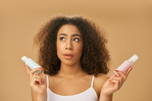 Attractive healthy african american young woman looking doubtful while choosing between two beauty products, posing isolated over beige background. Skincare concept