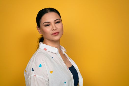 Charming Asian woman standing sideways looking at camera, wearing casual white shirt, isolated on yellow background. Human emotions, facial expression concept.