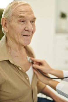 Close-up photo of an optimistic elderly citizen looking fine while being examined with a stethoscope