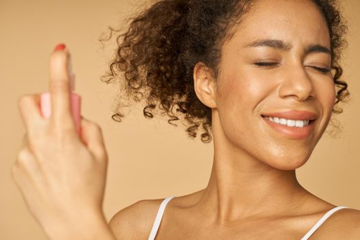 Close up portrait of happy young woman applying spray water on her face, posing isolated over beige background. Beauty, skincare routine concept