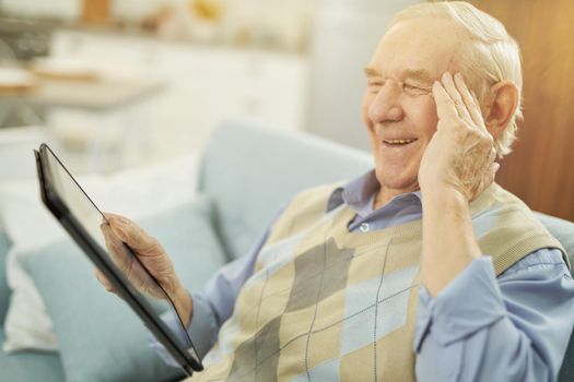 Smiling male pensioner holding gadget while looking at the tablet screen and making a video call with doctor