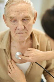 Tranquil elderly man looking a bit sad while having his lungs and heart checked by a doctor with stethoscope