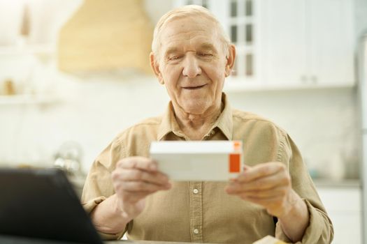 Calm senior citizen holding and looking at the tiny box of medication while sitting at the table at home