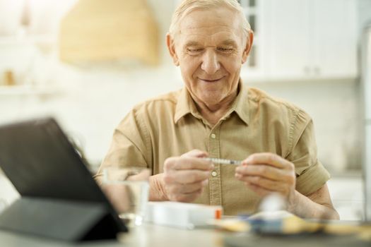 Joyous senior man smiling while sitting at the table and looking at a syringe in his hands