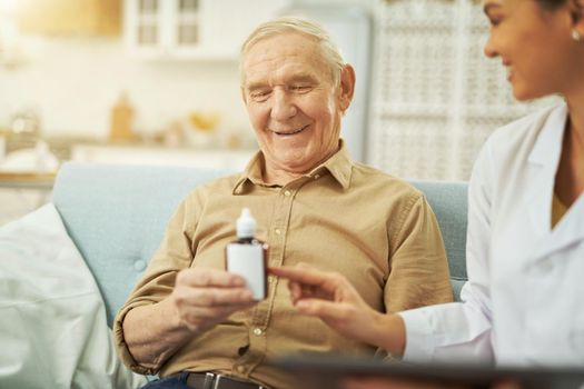 Happy nurse explaining to an elderly man how to take medicine while sitting on the couch at home