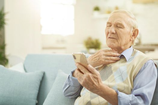 Gray-haired man sitting on the couch and explaining to the doctor that he has a sore throat while using phone