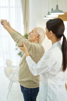 Side view of old man raising his hands up and doing exercises with fitness rubber band at home, nurse near him