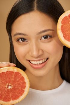 Close up portrait of joyful natural mixed race young woman smiling at camera, holding grapefruit cut in half, posing isolated over beige background. Health and beauty concept