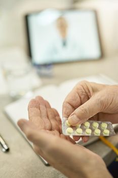 Selective focus photo of elderly person taking pills out of the blister while listening to doctor on a video-call