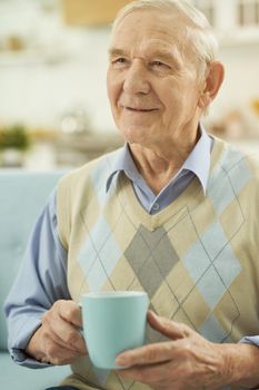 Happy old-aged man enjoying of hot drink while sitting in living room at home