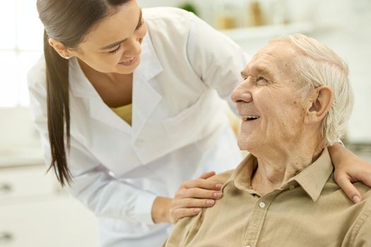 Contented elderly man smiling and looking at young medical doctor touching his shoulder
