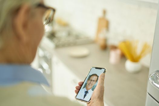 Cropped photo of grey-haired man in glasses holding a smartphone and video-calling his medical doctor