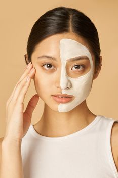 Portrait of young woman with facial mask applied on half of her face receiving spa treatments, posing isolated over beige background. Front view