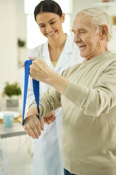 Happy pensioner doing exercises and holding a fitness elastic band in hands while talking to female doctor
