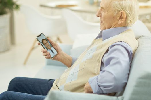 Side view of old man sitting on sofa at home and using mobile phone during a consultation with a therapist