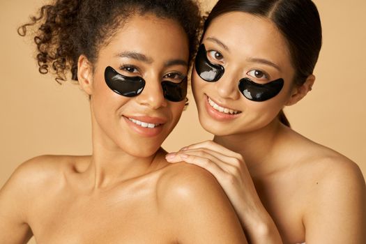 Beauty portrait of two adorable young women posing with applied black under eye patches, standing together isolated over beige background. Skincare routine concept