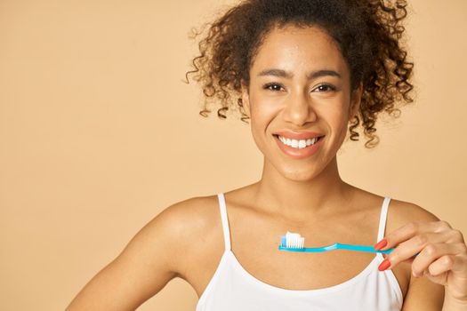 Portrait of cheerful young mixed race woman brushing teeth in the morning, posing isolated over beige background. Dental health care concept