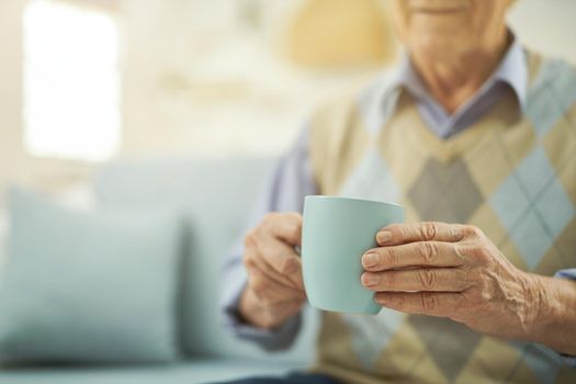 Cropped photo of old-aged man enjoying of hot drink while sitting in living room at home. Copy space