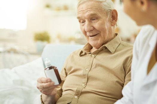 Senior citizen holding medicine and sitting near nurse in room. Copy space