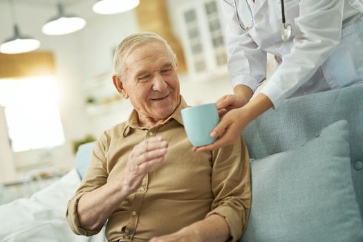 Caring female doctor in white coat serving a cup to senior citizen at his house while taking care of him