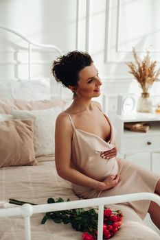 Lovely pregnant lady looking peaceful while sitting on the bed beside a bouquet of roses and caressing her stomach