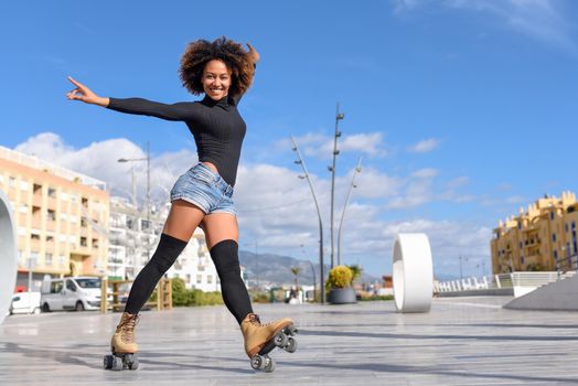 Young fit black woman on roller skates riding outdoors on urban street with open arms. Smiling girl with afro hairstyle rollerblading on sunny day