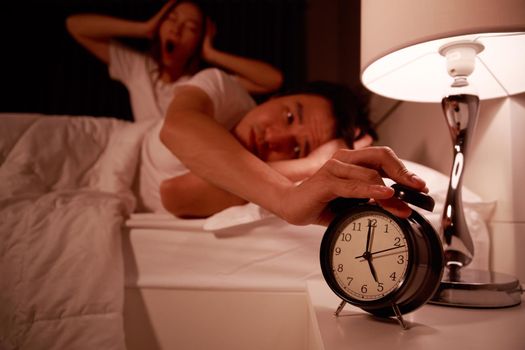 sleepy young couple in bed with extending hand to alarm clock in the morning