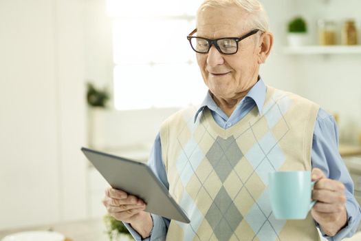 Elderly man in glasses holding a cup of tea while looking at the tablet screen and making a video call