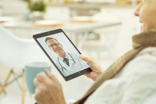 Elderly sick male holding a cup of drink while looking at the tablet screen and making a video call