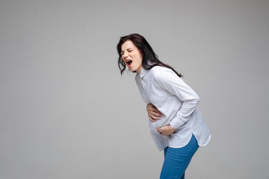 Portrait of crying and screaming Caucasian brunette in white shirt and jeans embracing her tummy and bending forward in pain. Cutout on grey background.