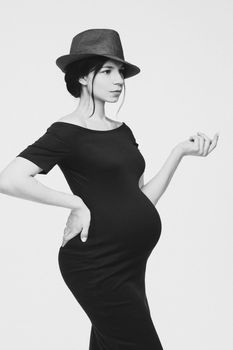 Black and white of gorgeous pregnant woman in black dress and hat posing on white background. Monochrome photo of fashionable elegant woman expecting a baby.