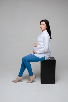 Full length studio portrait of attractive Caucasian brunette in white shirt and blue jeans and beige heels sitting on black cube embracing her belly and looking at camera.