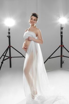 Full length of sensual cheerful attractive woman expecting a baby embracing her belly while posing with lights in background. Smiling pregnant woman with breast covered with white long cloth. Maternity concept.