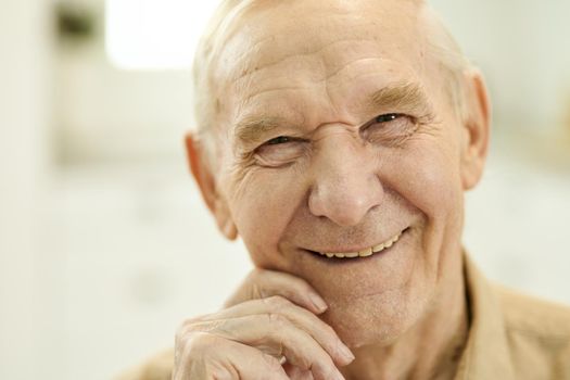 Close-up photo of a cheerful aged man smiling and touching his chin while looking at the camera