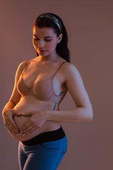 Studio portrait of attractive slim pregnant woman with dark hair with band standing in bra and jeans holding heart gesture with hands on tummy. Isolate on gentle pink background.