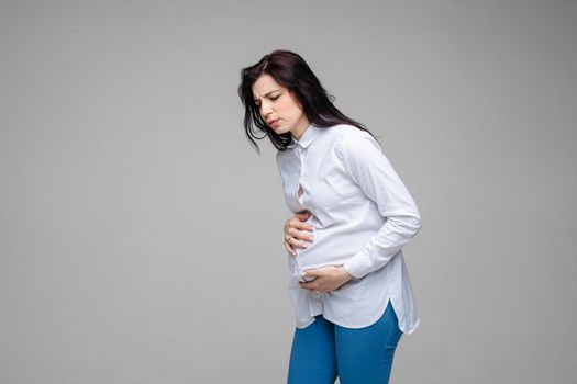 Pregnant brunette Caucasian woman feeling abdominal pain in her tummy feeling bad and making grimace. Isolate on grey in studio.