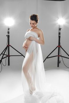 Full-length studio photo of brunette pregnant lady in a transparent white cloth posing in front of lamps