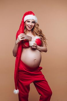 Smiling pregnant girl in Santa Claus suit eating gingerbread cookies with milk having fun full shot. Beautiful festive future mother celebrating Christmas holiday isolated at nude studio background