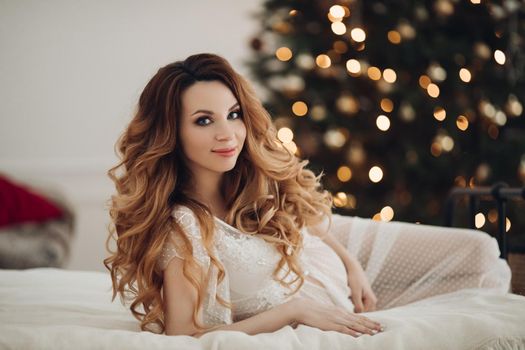 Beautiful smiling pregnant woman in white dress posing with New Year tree on background. Christmas and New Year concept