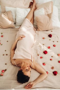 Upside-down shot of a relaxed brunette woman in beige dress laying on a bed sprinkled with flower petals