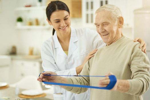 Smiling nurse in a white coat standing and explaining for male pensioner how to do exercises at home with fitness rubber