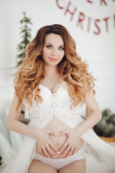 Portrait of smiling pregnant young woman tenderness holding big tummy showing heart medium shot. Beautiful girl making love gesturing posing enjoying pregnancy at Christmas tree background