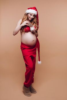 Smiling pregnant girl in Santa Claus suit eating gingerbread cookies with milk having fun full shot. Beautiful festive future mother celebrating Christmas holiday isolated at nude studio background