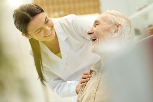 Blurred photo of an amicable medical doctor smiling at senior patient while touching his shouder