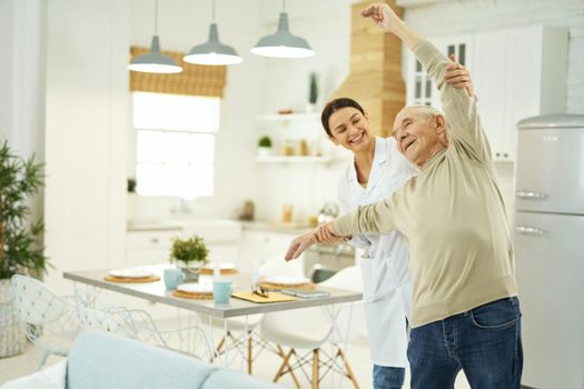 Smiling young female doctor in white coat helping an elderly man to do exercises in his apartment