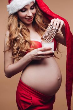 Smiling beautiful pregnant lady in Santa Claus hat soaking gingerbread in milk on beige background. Christmas and New Year concept