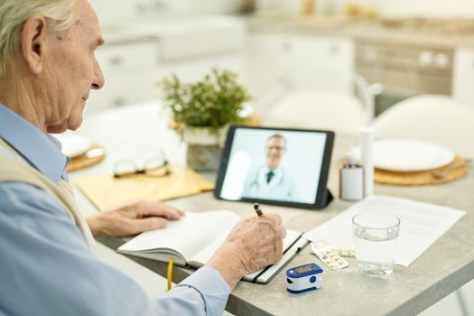 Responsible senior citizen taking notes while video-calling his doctor on a tablet from home